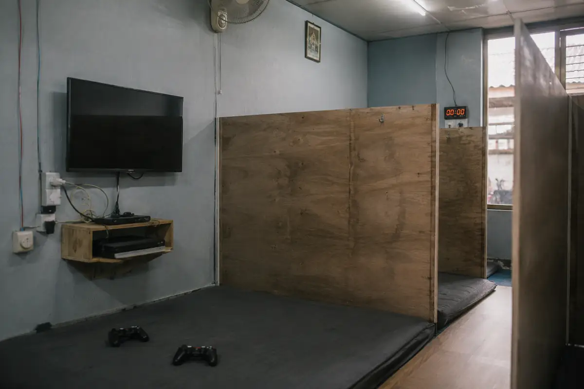 Bilik rental PlayStation dengan televisi di dinding dan konsol di rak kayu.